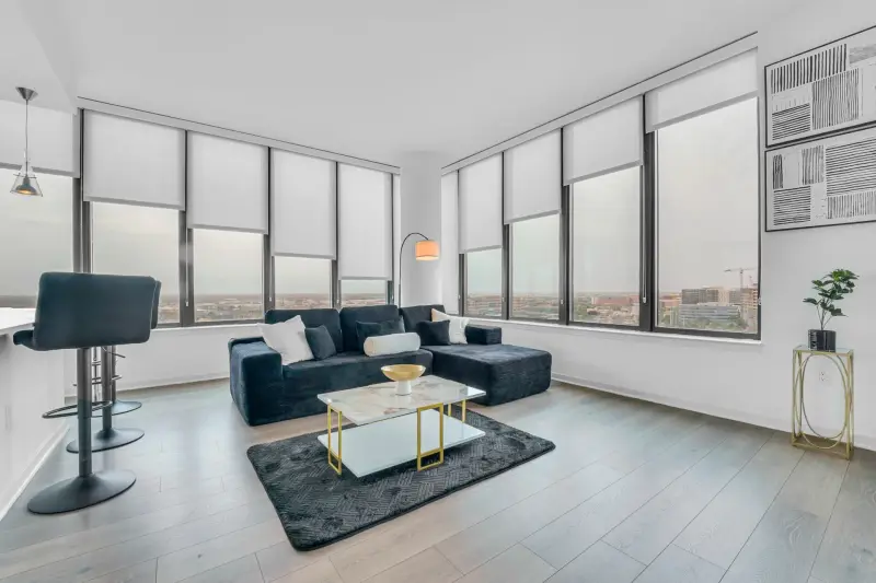 Sleek high-rise condo living room wrapped in floor-to-ceiling windows with a city skyline view, navy sectional and brass accents. Distinctive urban contrast to suburban frames. — Houston real estate photography
