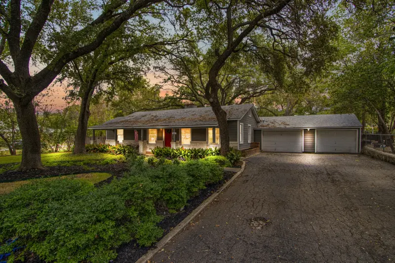 Ranch home framed by mature live oaks at dusk, warm interior glow and red front door under a soft pastel sky. — San Antonio real estate photography