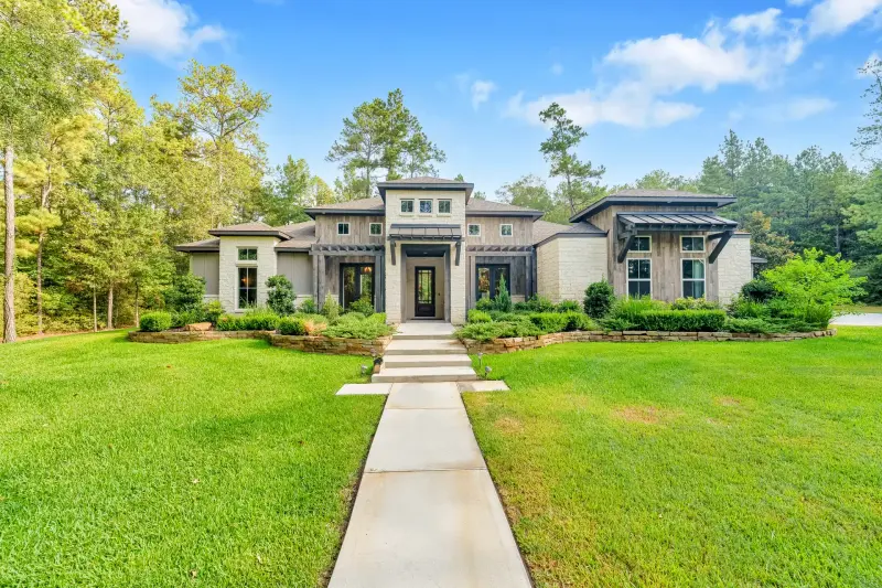 Modern stone-and-wood luxury home, dead-center symmetric concrete walkway leading to entry, lush manicured beds framed by tall pines, clean blue sky, straight verticals, tack sharp. Hero-grade exterior. — Houston real estate photography
