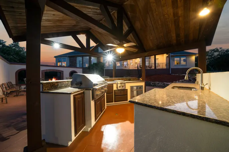 Twilight outdoor kitchen under a timber pavilion; fireplace and home aglow. — Austin–San Antonio corridor real estate photography